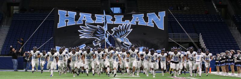Football team running on the field with the Harlan banner in the background.